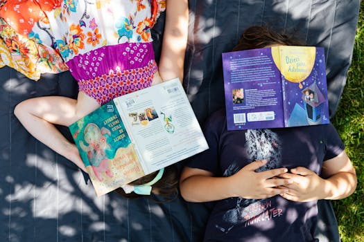 Two children lie outdoors on grass, faces covered by books, enjoying a sunny day.